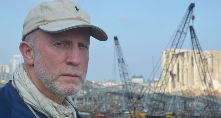 Andy Kent stares solemnly at the camera, behind him is debris from destroyed buildings and construction equipment.