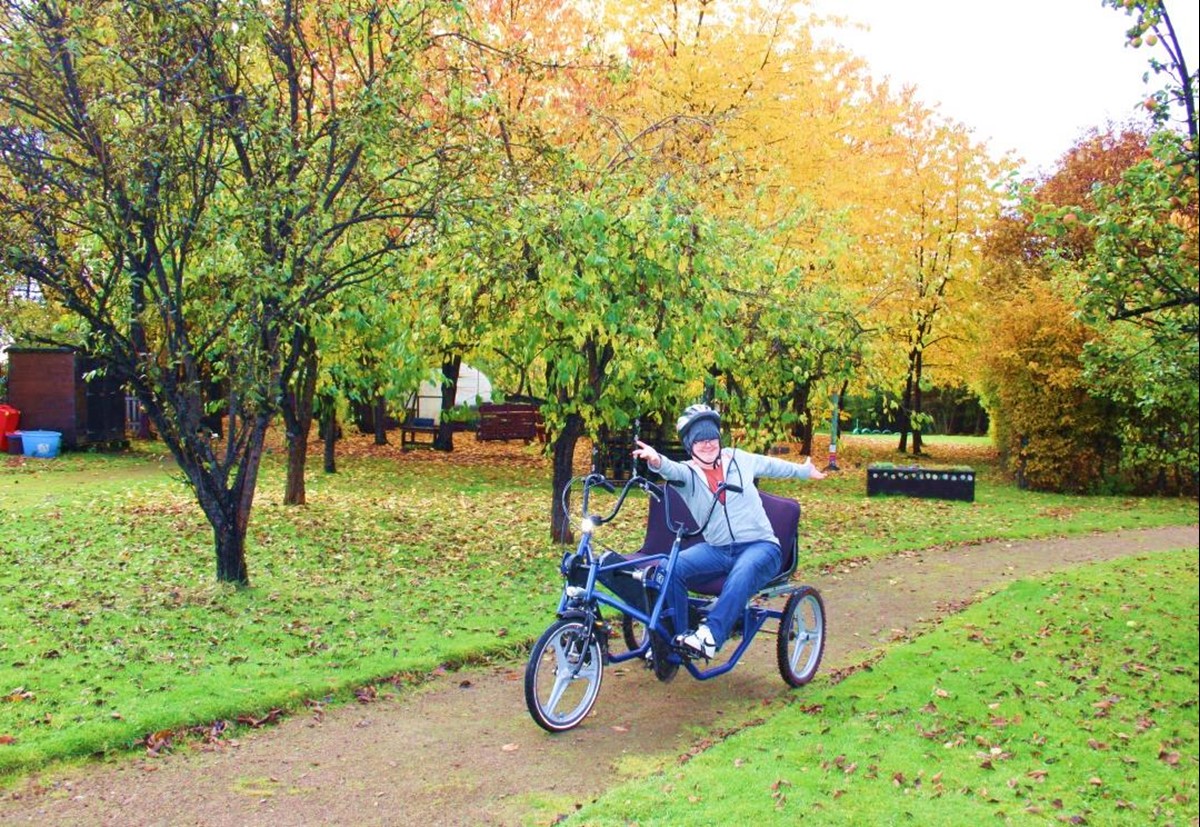 A supported person enjoying time on a tricycle outside. They are stretching their arms out happily. The leaves on the trees are orange and brown, and the grass it littered with leaves.