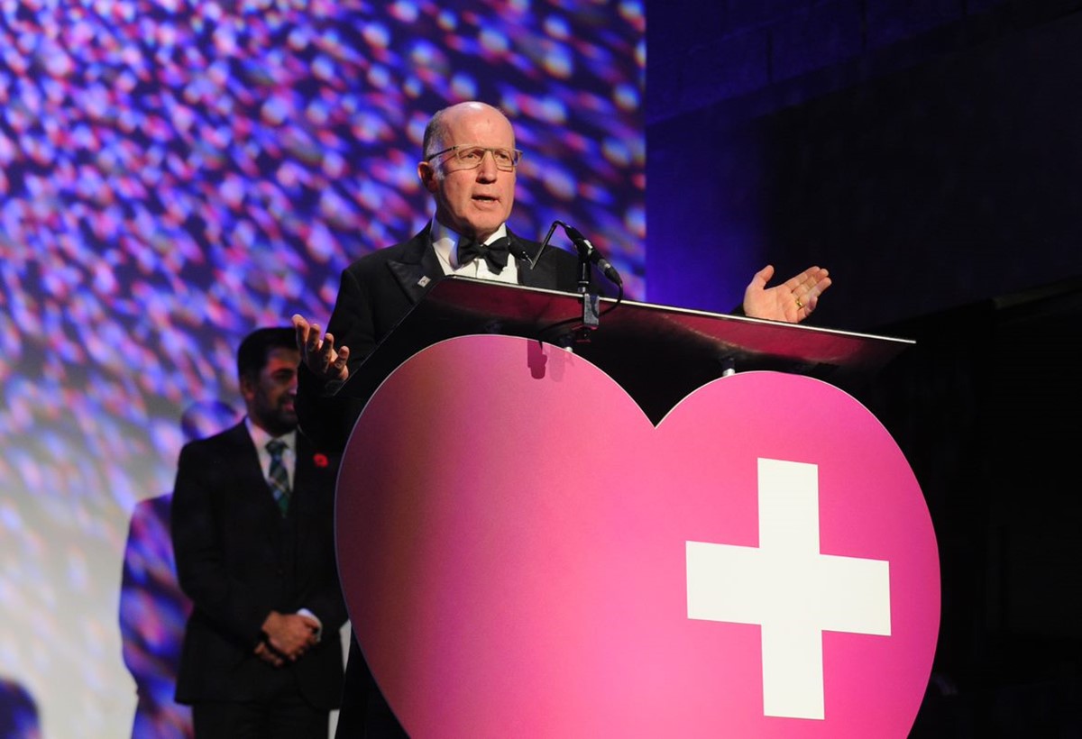 Mr.Kent, 58 years old, stands on stage behind a large pink love heart with a small white cross in the top right corner, the logo for the Scottish Health Awards. He is giving a speech after winning his award.